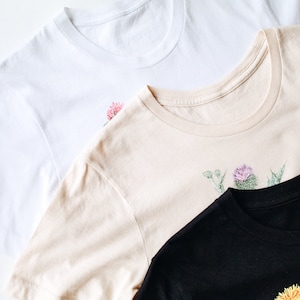 May include: Three t-shirts with floral designs. The top shirt is white with a pink flower, the middle shirt is beige with a purple flower, and the bottom shirt is black with a yellow flower.