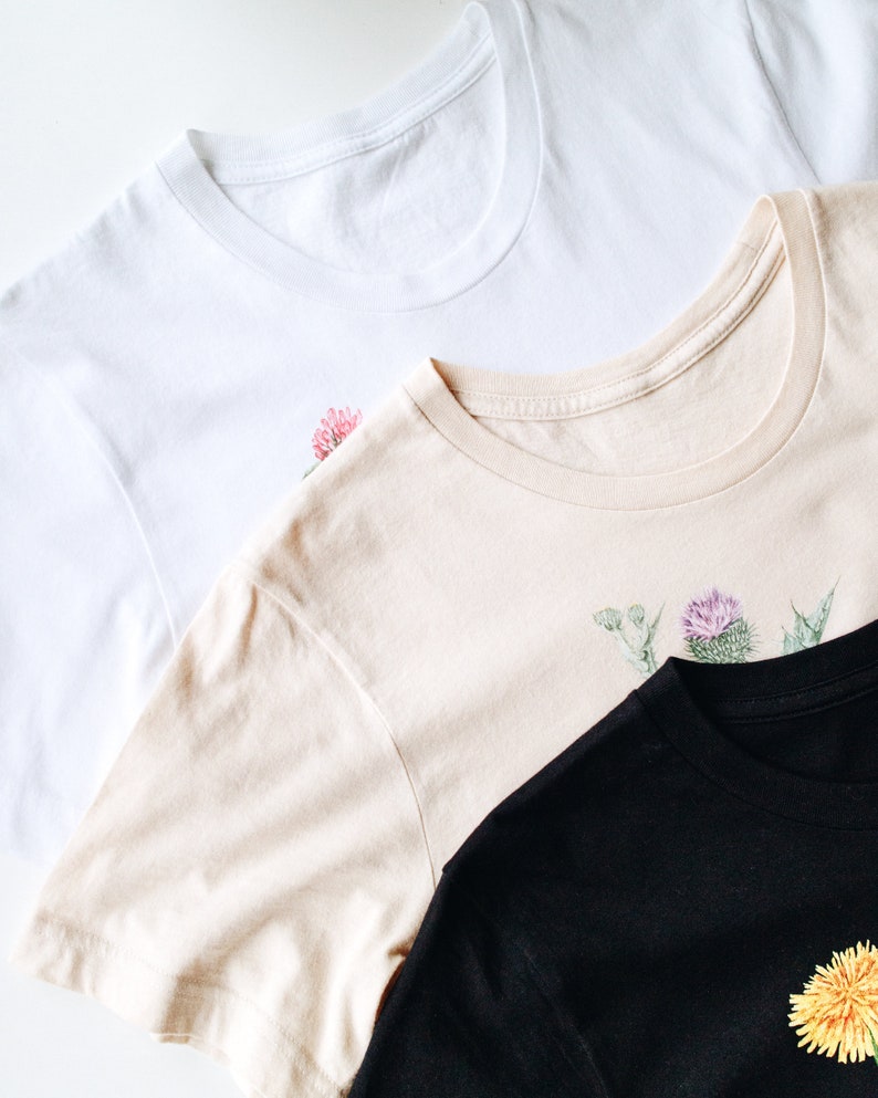 May include: Three t-shirts with floral designs. The top shirt is white with a pink flower, the middle shirt is beige with a purple flower, and the bottom shirt is black with a yellow flower.