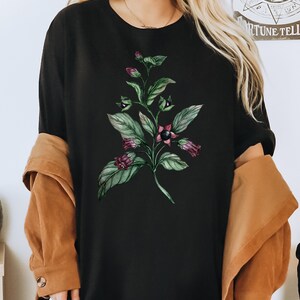 以下が含まれることがあります： 緑と紫の花柄がプリントされた黒いTシャツ。デザインは葉と花がついた植物です。