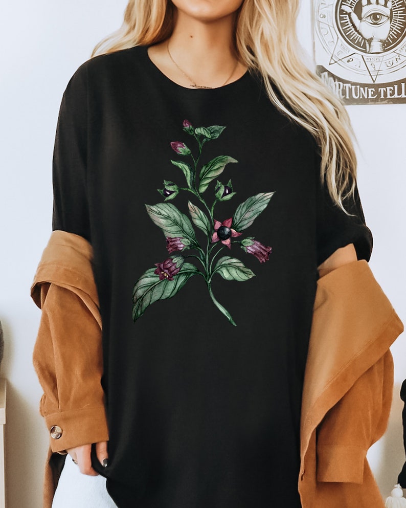 以下が含まれることがあります： 緑と紫の花柄がプリントされた黒いTシャツ。デザインは葉と花がついた植物です。