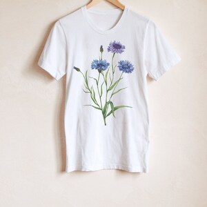 May include: White t-shirt with a floral design featuring blue and purple flowers with green stems and leaves.