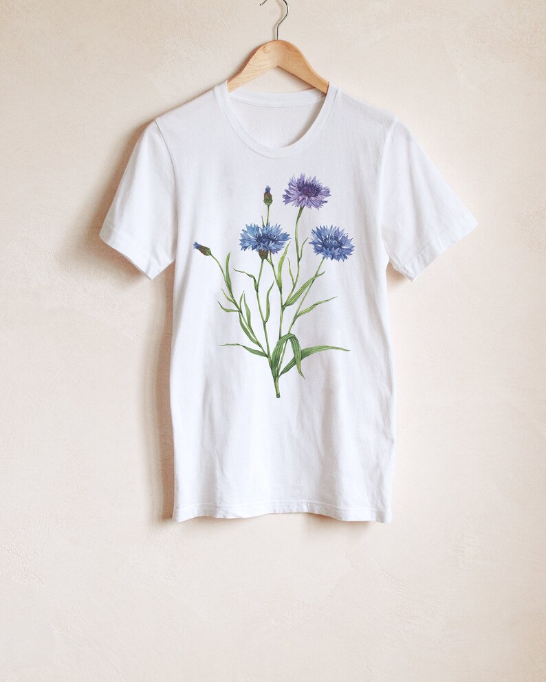 May include: White t-shirt with a floral design featuring blue and purple flowers with green stems and leaves.