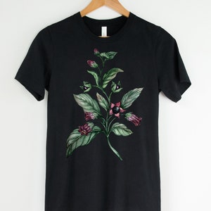 以下が含まれることがあります： 黒いTシャツに、緑の葉と紫の花、黒い実がついた植物の水彩画が描かれています。