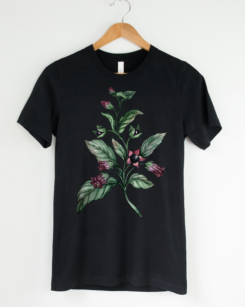 以下が含まれることがあります： 黒いTシャツに、緑の葉と紫の花、黒い実がついた植物の水彩画が描かれています。