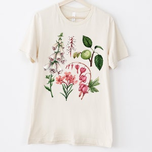 May include: A beige t-shirt with a floral print featuring white foxgloves, pink bleeding hearts, and green leaves.