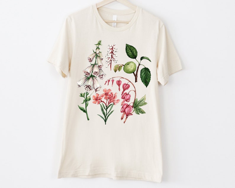 May include: A beige t-shirt with a floral print featuring white foxgloves, pink bleeding hearts, and green leaves.