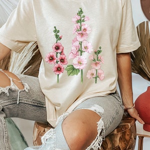 May include: A beige t-shirt with a floral print of pink and white flowers. The flowers are arranged in a vertical line and have green stems and leaves.
