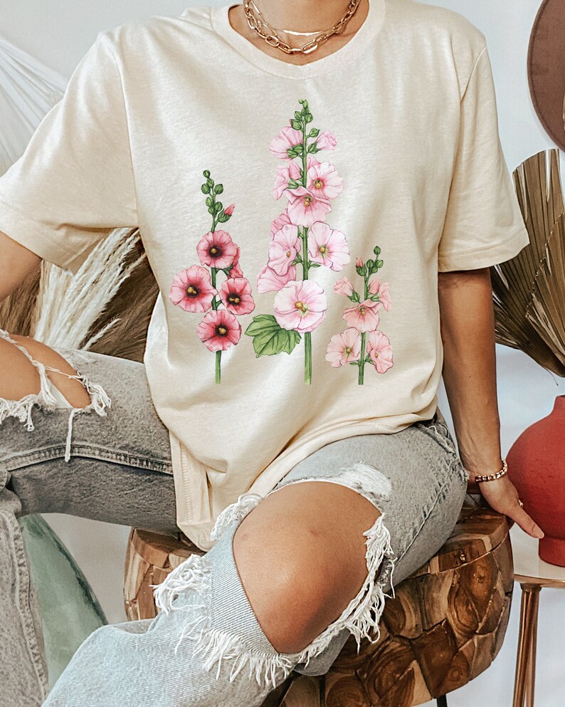 May include: A beige t-shirt with a floral print of pink and white flowers. The flowers are arranged in a vertical line and have green stems and leaves.