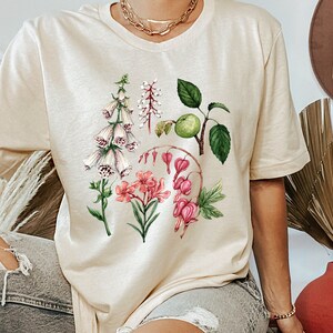 May include: A beige t-shirt with a floral print featuring pink, white, and green flowers. The flowers are arranged in a cluster and include foxgloves, bleeding hearts, and apple blossoms.