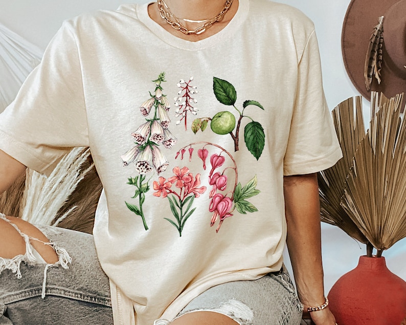 May include: A beige t-shirt with a floral print featuring pink, white, and green flowers. The flowers are arranged in a cluster and include foxgloves, bleeding hearts, and apple blossoms.
