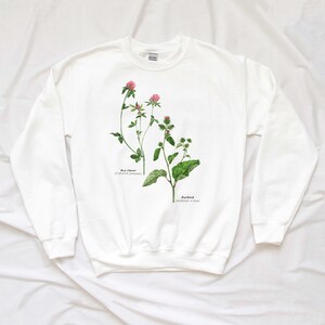 May include: White sweatshirt with a botanical illustration of red clover and burdock plants. The illustration is in watercolor style and includes the scientific names of the plants.