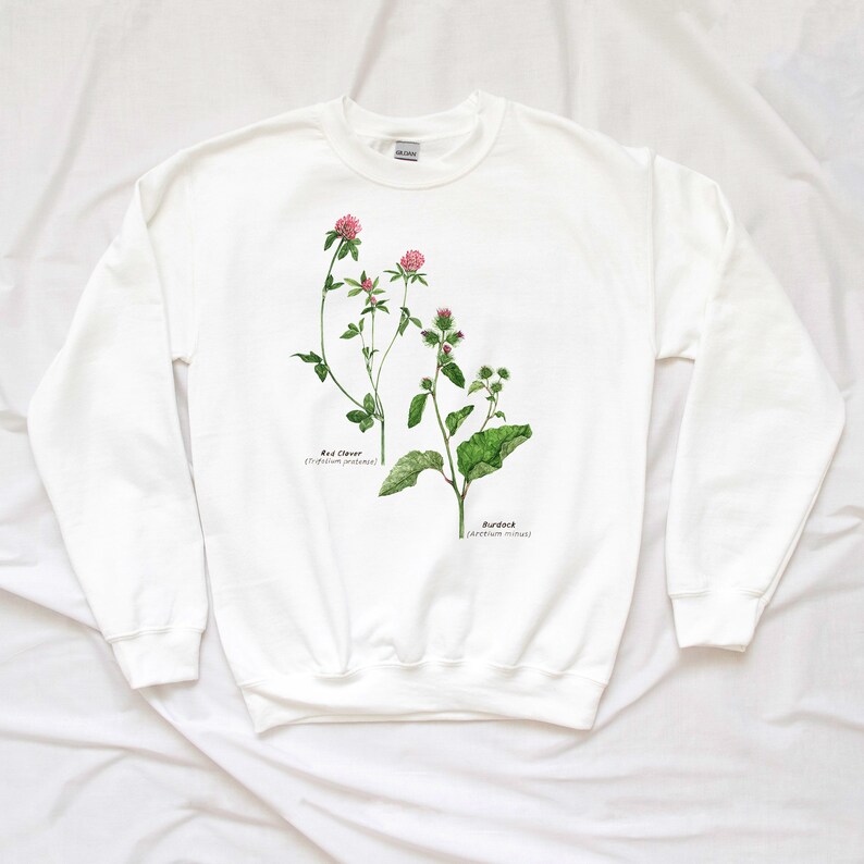 May include: White sweatshirt with a botanical illustration of red clover and burdock plants. The illustration is in watercolor style and includes the scientific names of the plants.