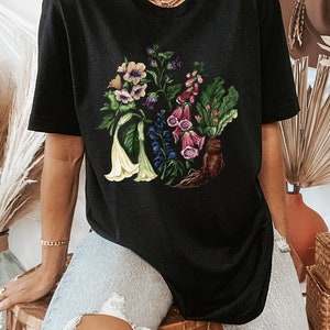 May include: Black oversized t-shirt with a floral print featuring a variety of flowers in shades of pink, purple, white, and green. The flowers are arranged in a circular pattern and have a vintage botanical illustration style.