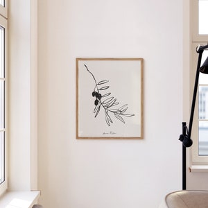 May include: A black and white line drawing of an olive branch with olives, framed in a light wood frame. The print is on white paper and has the artist's signature in the bottom right corner.