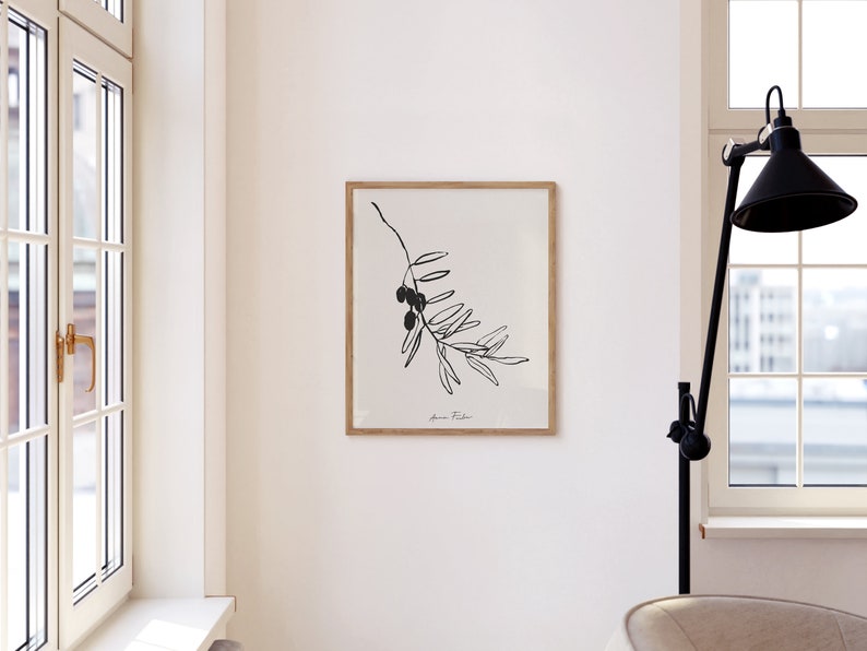 May include: A black and white line drawing of an olive branch with olives, framed in a light wood frame. The print is on white paper and has the artist's signature in the bottom right corner.