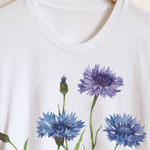 May include: White t-shirt with a floral design featuring three blue cornflowers with green stems and leaves.