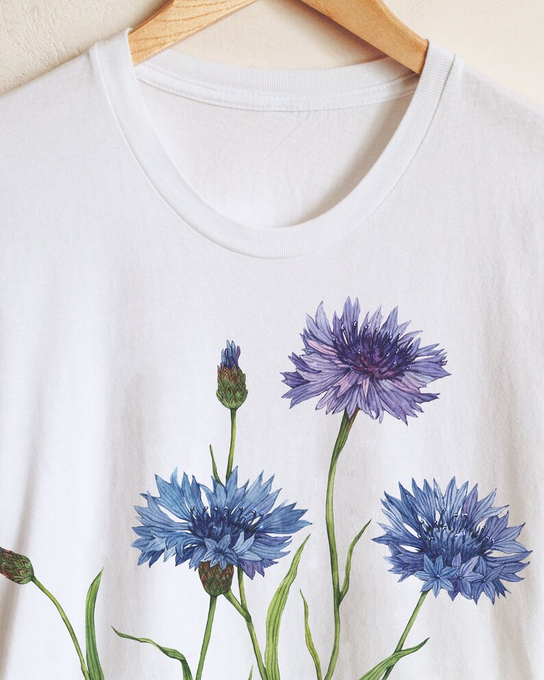 May include: White t-shirt with a floral design featuring three blue cornflowers with green stems and leaves.