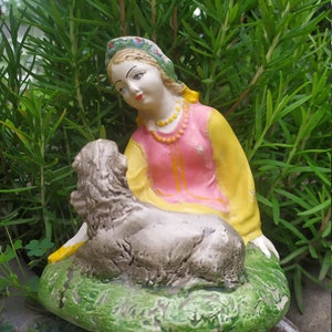 Könnte beinhalten: Eine Keramikfigur einer Frau in einem rosa und gelben Kleid und einem Blumenkopfband, die ein Lamm in ihren Armen hält. Die Figur sitzt auf einem grünen Sockel mit einem braunen Stein dahinter.