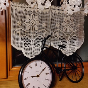 May include: A black metal bicycle clock with a white face and black Roman numeral hour markers. The clock face says "London 1866". The clock is sitting on a wooden surface with a white crocheted lace curtain hanging above it.