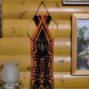 Op de afbeelding: Een macramé wandhanger met een ronde houten ring en zwart en oranje garen. De hanger heeft een geometrisch patroon en franjes.