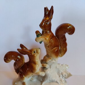 Może przedstawiać: Ceramiczna figurka przedstawiająca dwa wiewiórki. Jedna wiewiórka stoi, trzymając orzech, podczas gdy druga wydaje się skakać. Wiewiórki są brązowe z białymi detalami, na białej podstawie.
