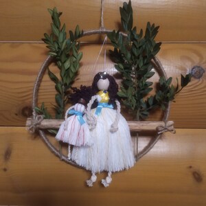 Pode incluir: Duas bonecas de fio com longos vestidos brancos e fitas azuis estão sentadas em um galho de madeira. As bonecas estão penduradas em um aro de macramê com folhagem verde.