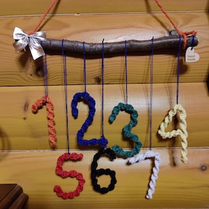 Op de afbeelding: Een houten tak met zeven gehaakte cijfers die eraan hangen. De cijfers zijn in verschillende kleuren: oranje, blauw, groen, geel, rood, zwart en wit. Een zilveren strik is aan de tak bevestigd. Een klein houten hartvormig label met de tekst "hand made" is aan de tak bevestigd.