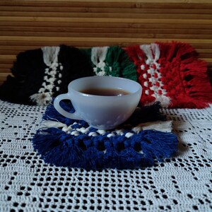 Könnte beinhalten: Eine weiße Keramik-Teetasse gefüllt mit Tee steht auf einem blauen und weißen gehäkelten Untersetzer. Der Untersetzer liegt auf einer weißen gehäkelten Tischdecke. Im Hintergrund befinden sich drei bunte gehäkelte Untersetzer.