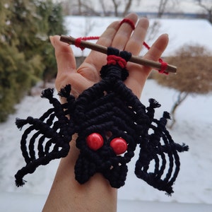 May include: Black macrame bat hanging ornament with red beads for eyes. The bat is hanging from a stick with red string.