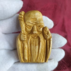 May include: A carved wooden figurine of an elderly man with a long beard, holding a staff. The figure is light brown and has detailed facial features. The carving is held in a white gloved hand against a red background.