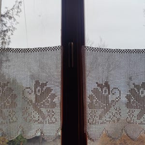 May include: Two white crocheted curtains with floral designs hang in a window. The curtains are made of a delicate, lacy material and have a scalloped edge.