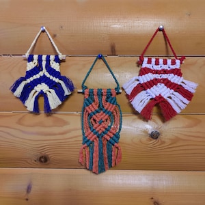 Op de afbeelding: Drie macramé wandhangers met verschillende kleurpatronen. De eerste is blauw en wit, de tweede is rood en wit en de derde is groen, oranje en wit.