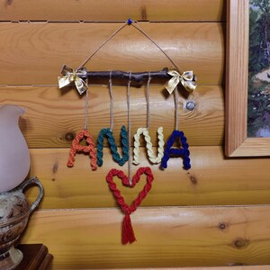 Op de afbeelding: Een kleurrijk garennaamschild met de naam "ANNA" gespeld in regenboogkleuren. De letters zijn bevestigd aan een klein takje met gouden strikken aan beide uiteinden. Een rood hart met een kwast hangt onder de letters.