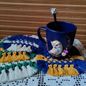 Könnte beinhalten: Eine marineblaue Tasse mit Eulenmotiv und ein dekorativer Löffel ruhen auf einem Makramee-Untersetzer. Der Untersetzer hat blaue, gelbe, grüne und weiße Quasten. Der Hintergrund ist eine Bambusmatte.