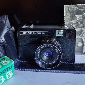 Puede incluir: Una cámara de película de 35 mm negra y plateada con una lente etiquetada como "TRIPLET 69-3 4/40" y el texto "вилия - VILIA" en la parte superior. La cámara está sobre una superficie blanca.