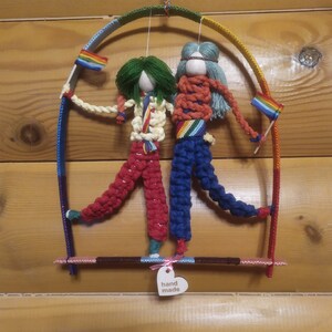 May include: Handmade macrame art piece featuring two figures on a swing. The figures have yarn hair, one green and one blue, and wear colorful outfits. Each figure holds a small rainbow flag. The swing is suspended from a rainbow-colored arch.