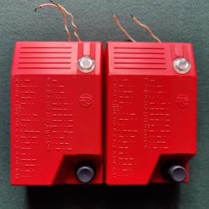 Può includere: Due dispositivi in plastica rossa con un alfabeto braille e numeri stampati sul davanti. Ogni dispositivo ha una piccola manopola nera e un pulsante bianco sulla parte superiore.