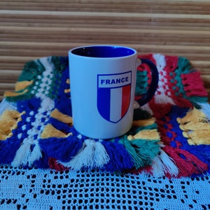 Könnte beinhalten: Weiße Keramiktasse mit blauem Innenraum und Henkel. Die Tasse zeigt ein blaues, weißes und rotes Schild mit dem Wort "FRANCE" darüber.