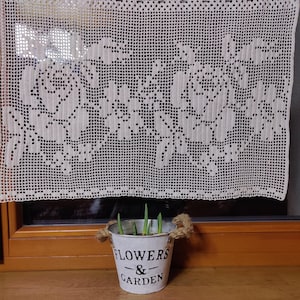 May include: A white metal flower pot with the text "FLOWERS & GARDEN" on the side. The pot is filled with green plant sprouts and has a brown rope handle.
