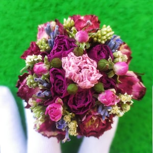 Könnte beinhalten: Ein Strauß getrockneter Blumen in Rosa-, Lila- und Grüntönen. Die Blumen sind kreisförmig angeordnet und mit einem weißen Band zusammengehalten.