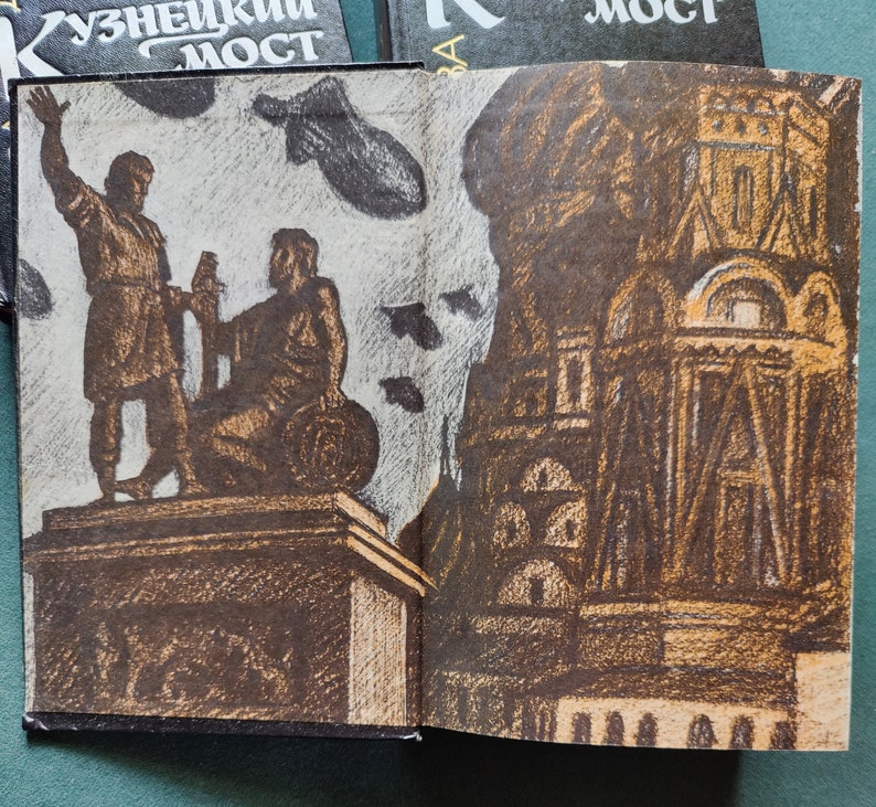 May include: An open book with a brown and white illustration of a statue of two figures standing on a pedestal. The background of the illustration is a light gray with black birds flying. The book is open to a page with a brown and white illustration of a building with a large window. The book cover has the Russian text 'Кузнецкий Мост' which translates to 'Kuznetsky Most'.