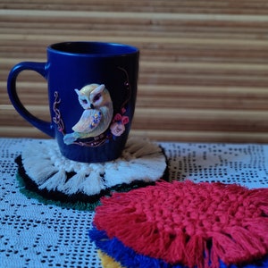 Könnte beinhalten: Eine blaue Keramiktasse mit einem weißen Eulenmotiv und rosa Blumen. Die Tasse steht auf einem weißen gehäkelten Untersetzer mit Fransen. Im Vordergrund befindet sich ein roter, blauer und gelber gehäkelter Untersetzer mit Fransen.