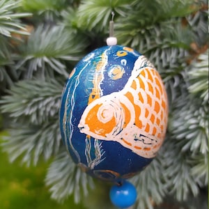 May include: A blue and orange hand-painted egg ornament with a fish design. The egg is hanging from a white string and has a blue bead at the bottom.
