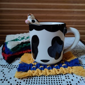 Könnte beinhalten: Eine weiße Keramiktasse mit schwarzem Rand und schwarzen Kuhflecken. Die Tasse hat einen schwarzen Henkel und eine kleine Kuhfigur am Rand. Die Tasse steht auf einem gelben und blauen gehäkelten Untersetzer.