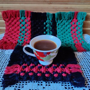 Könnte beinhalten: Eine Tasse Kaffee auf einem gewebten Untersetzer in Schwarz, Rot und Grün. Der Untersetzer liegt auf einer weißen gehäkelten Oberfläche.