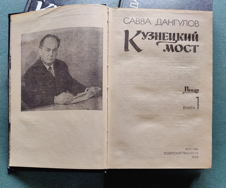 May include: A book cover with a black and white photo of a man in a suit. The book title is 'Кузнецкий Мост' in Russian. The author is 'Савва Дангулов' in Russian. The book is a novel and is the first book in a series. The book was published in Moscow by 'Советский Писатель' in 1980.