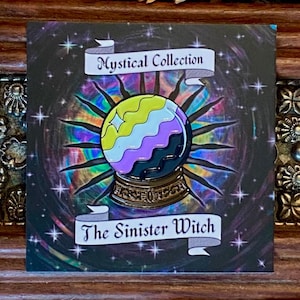 May include: A black and gold enamel pin featuring a crystal ball with a yellow, white and purple striped design. The pin is set against a black background with a rainbow swirl and stars. The text "Mystical Collection" and "The Sinister Witch" are displayed on banners.
