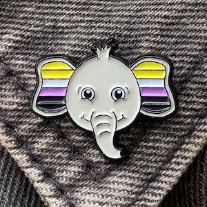 Könnte beinhalten: Eine weiße Elefanten-Anstecknadel mit einem gelben, weißen, violetten und schwarzen Streifenmuster an den Ohren. Die Anstecknadel ist ein Elefant im Cartoon-Stil mit einem lächelnden Gesicht und einem kleinen Rüssel.