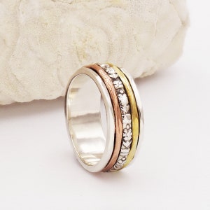 Può includere: Un anello girevole in argento con tre bande di rame, ottone e argento. Le bande sono decorate con piccoli disegni in rilievo.