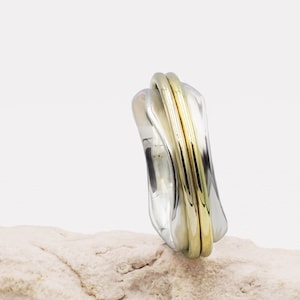 Può includere: Un anello in argento e oro con un designo unico e ondulato. L'anello è composto da due bande, una in argento e una in oro, che sono intrecciate.